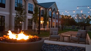 A fire pit in a landscaped area with outdoor furniture, string lights, and a house in the background. - Hotel Renovo Des Moines Urbandale,Tapestry Collection Hilton (Urbandale)