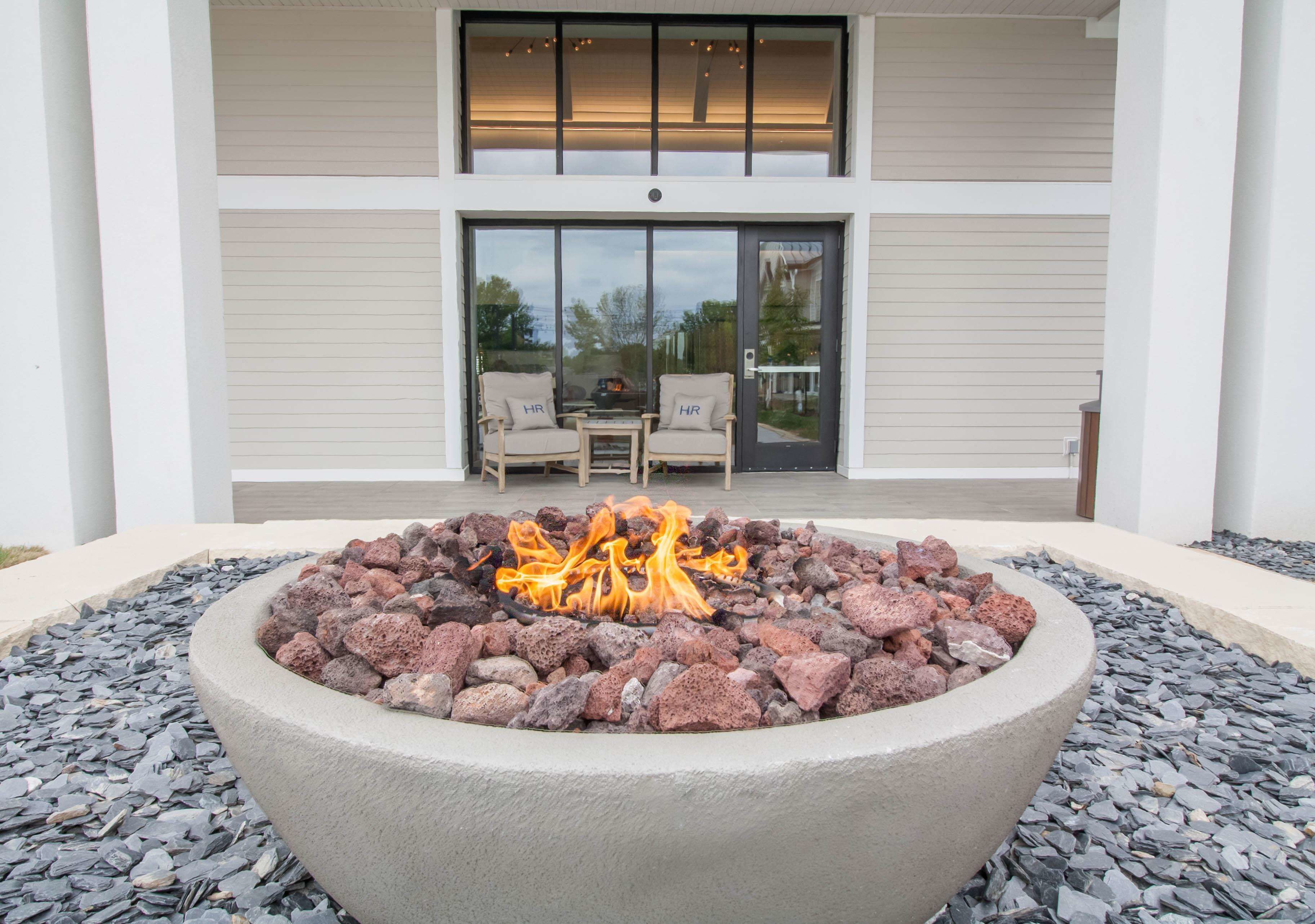 A fire pit with rocks in the center, surrounded by a stone border, in front of a building with a sliding glass door and two chairs outside.