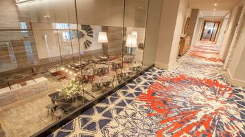 Un hall de hotel con alfombra estampada, paredes de vidrio y una zona de estar con mesas y sillas.
