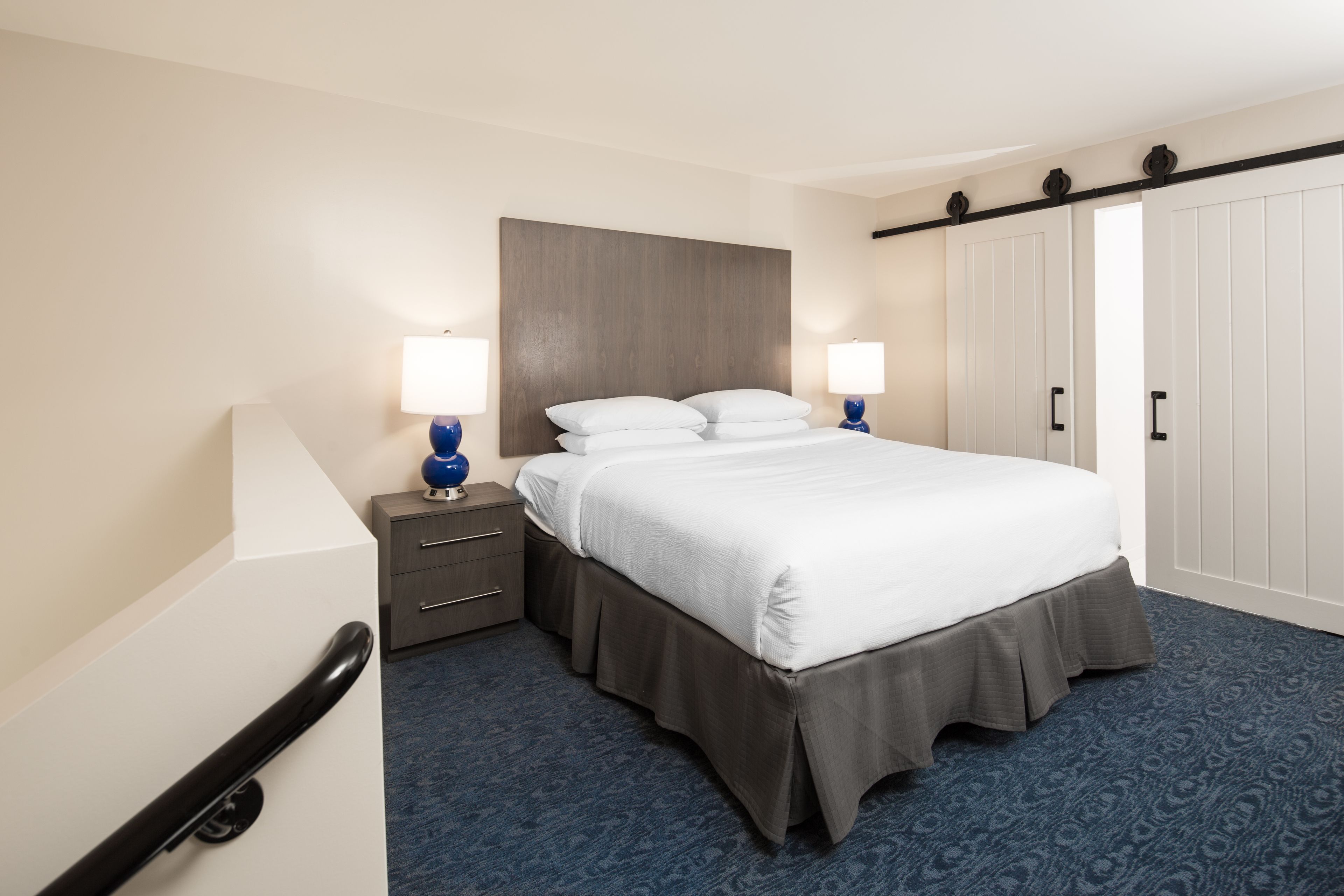 Una habitación de hotel con cama, mesitas de noche, lámparas, un armario y una alfombra azul.