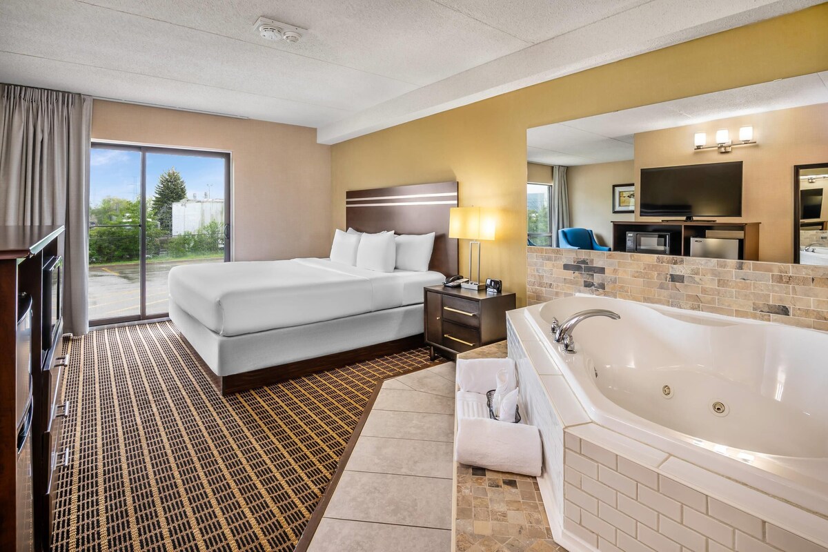 A hotel room with a bed, a large window, a TV, a sofa, a bathroom with a bathtub, and a mirror.