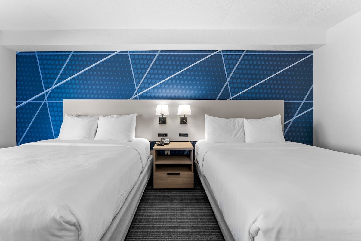 A hotel room with two beds, a nightstand with a lamp, and a blue patterned wall.