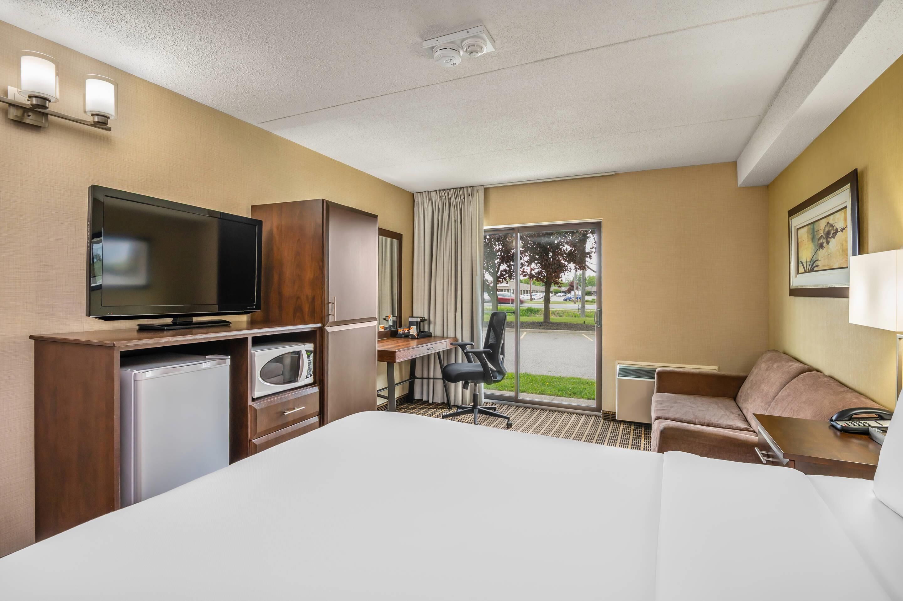 Een hotelkamer met een bed, een bureau met stoel, een televisie, een koelkast en een raam met uitzicht op de omgeving.
