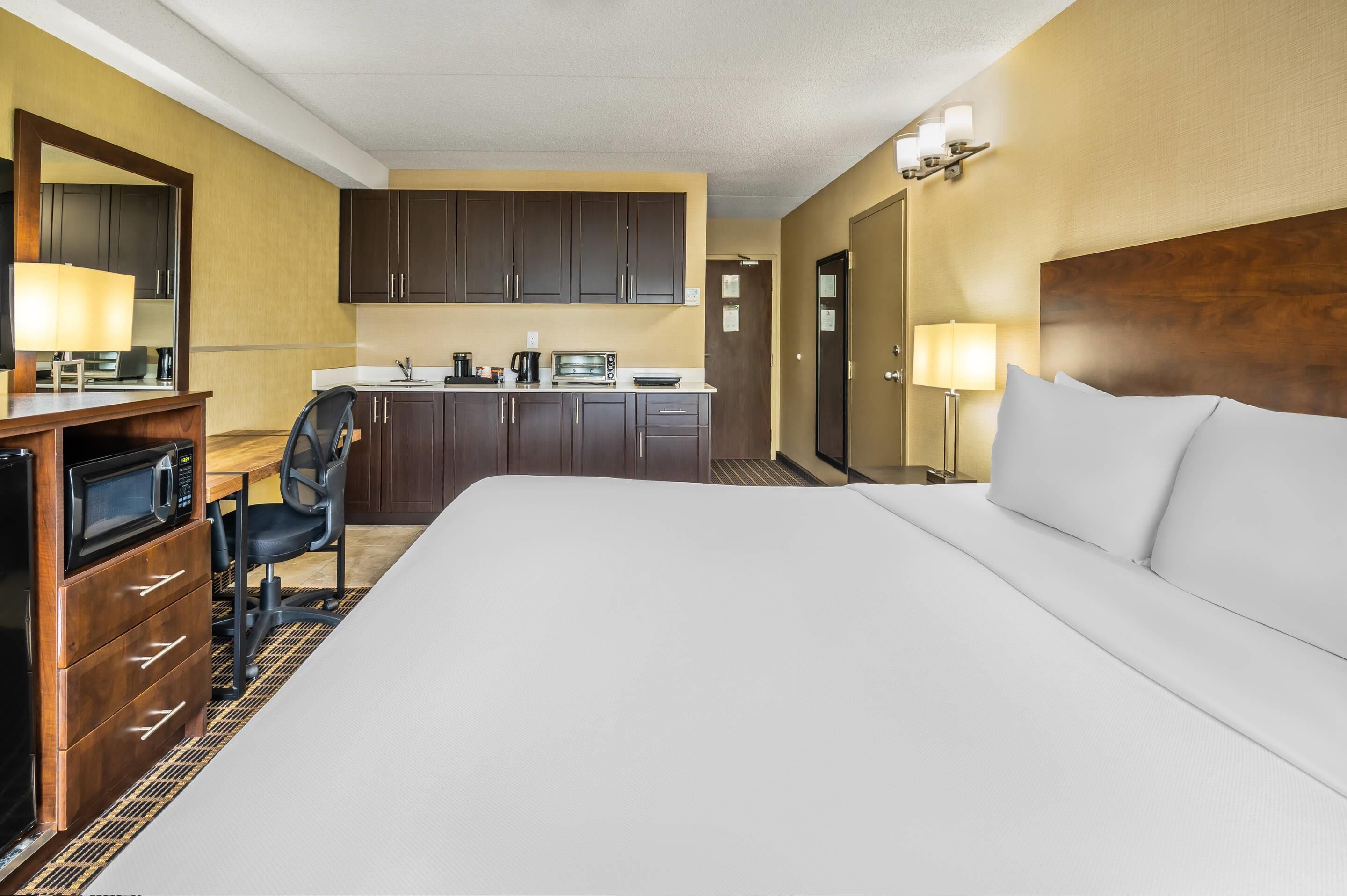 A hotel room with a large bed, a desk with a microwave, a chair, a kitchenette, and a door leading to another room.