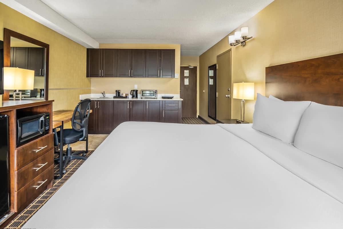A hotel room with a large bed, a desk with a microwave, a chair, a kitchenette, and a door leading to another room.