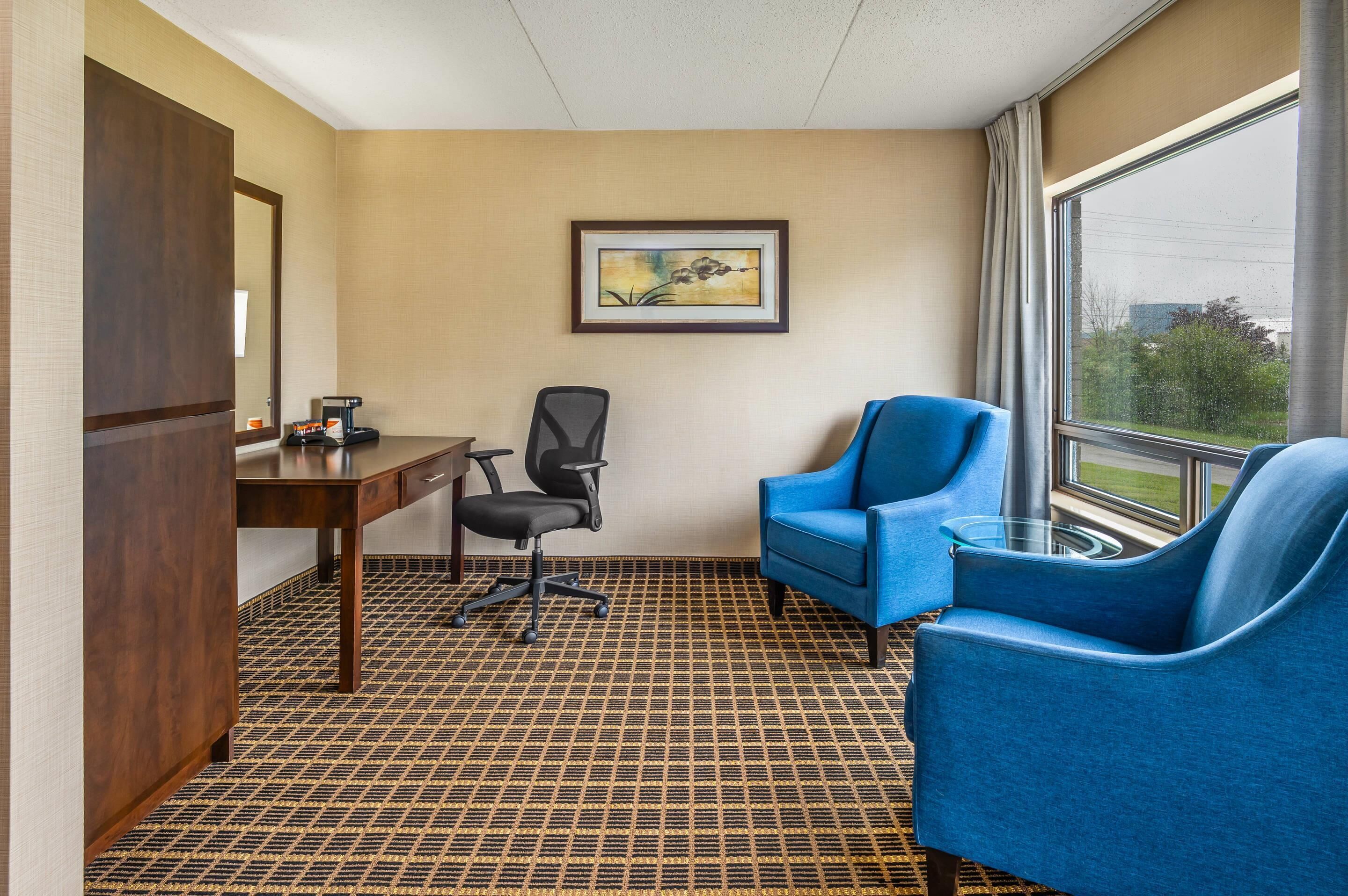 Habitación de hotel con un escritorio de madera, una silla de oficina negra, un sillón azul, una mesa de centro de vidrio y un ventanal con vistas a zonas verdes.