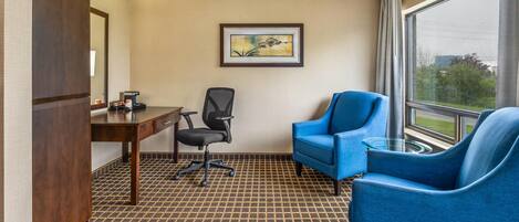 Habitación de hotel con un escritorio de madera, una silla de oficina negra, un sillón azul, una mesa de centro de vidrio y un ventanal con vistas a zonas verdes.