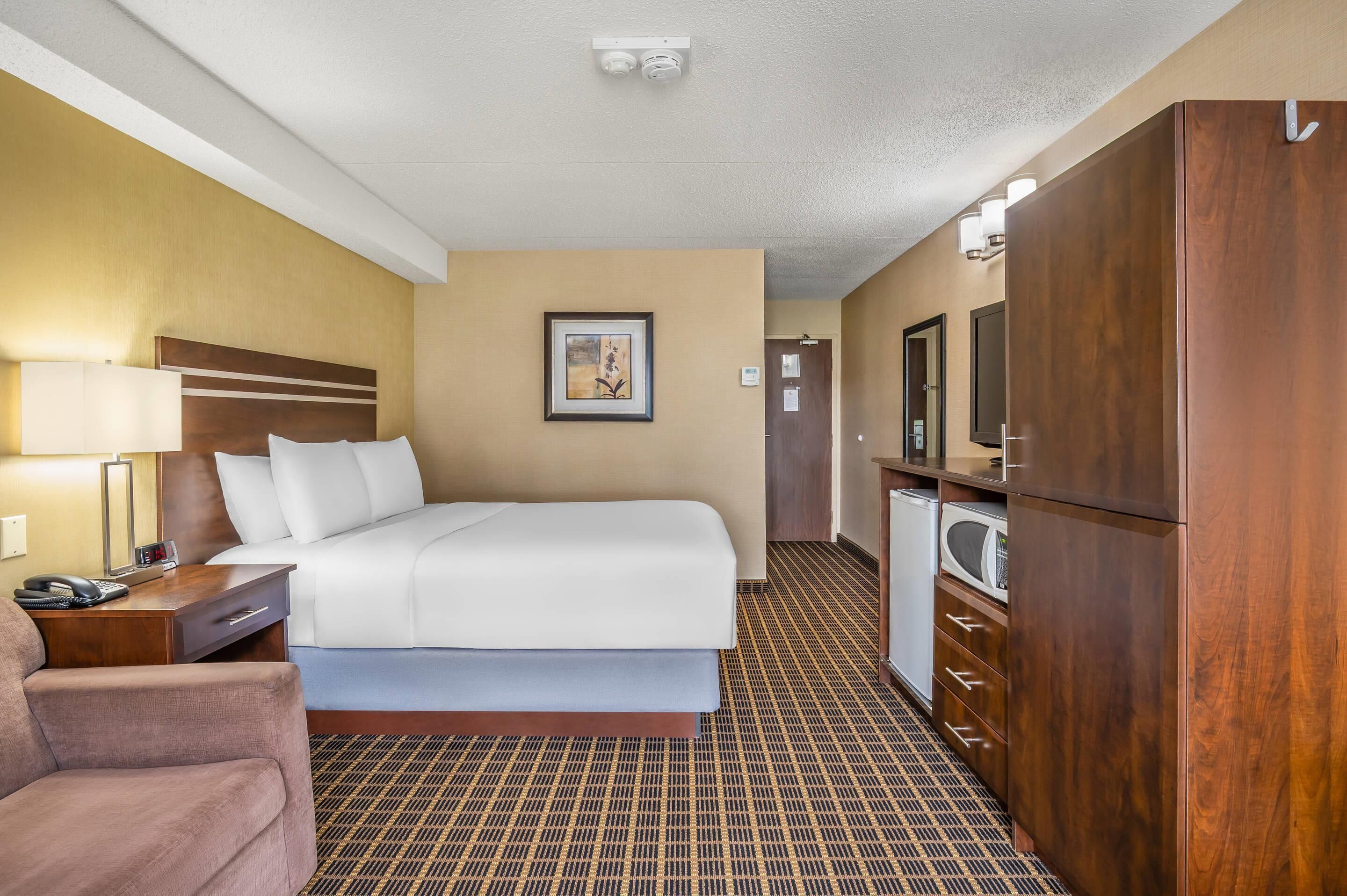 A hotel room with a bed, a desk with a lamp, a chair, a wardrobe, and a mirror.