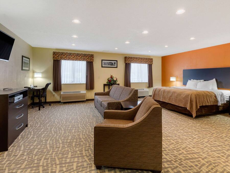 Habitación de hotel con cama, dos sillones, un escritorio con silla, televisor y dos ventanas con cortinas.
