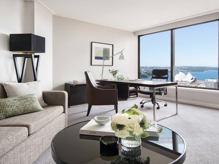 Une chambre d’hôtel moderne équipée d’un canapé, d’un bureau, d’une chaise, d’une table basse avec un livre et des fleurs, et d’une grande fenêtre donnant sur la ville.
