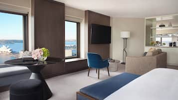 A hotel room with a bed, a blue chair, a small table with a vase of flowers, a flat-screen TV, and a view of a cityscape.