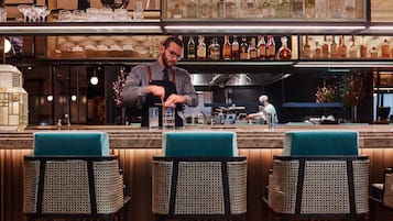 Eine Barbereich mit einem Bartender, der Getränke zubereitet, einer Küche im Hintergrund und Barhocker mit gewobenen Sitzflächen.
