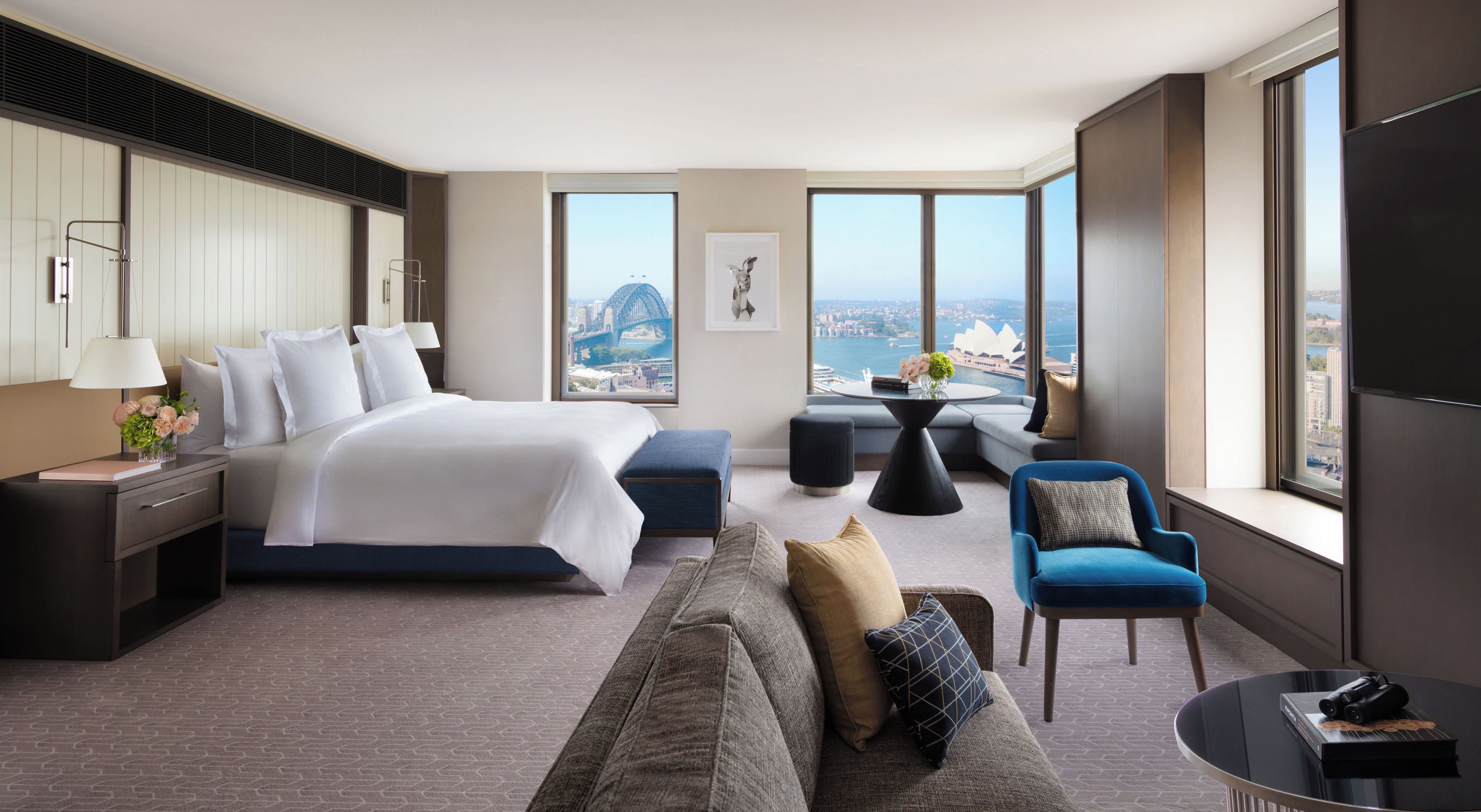 A modern hotel room with a large bed, a sofa, a small table, and a view of a cityscape.