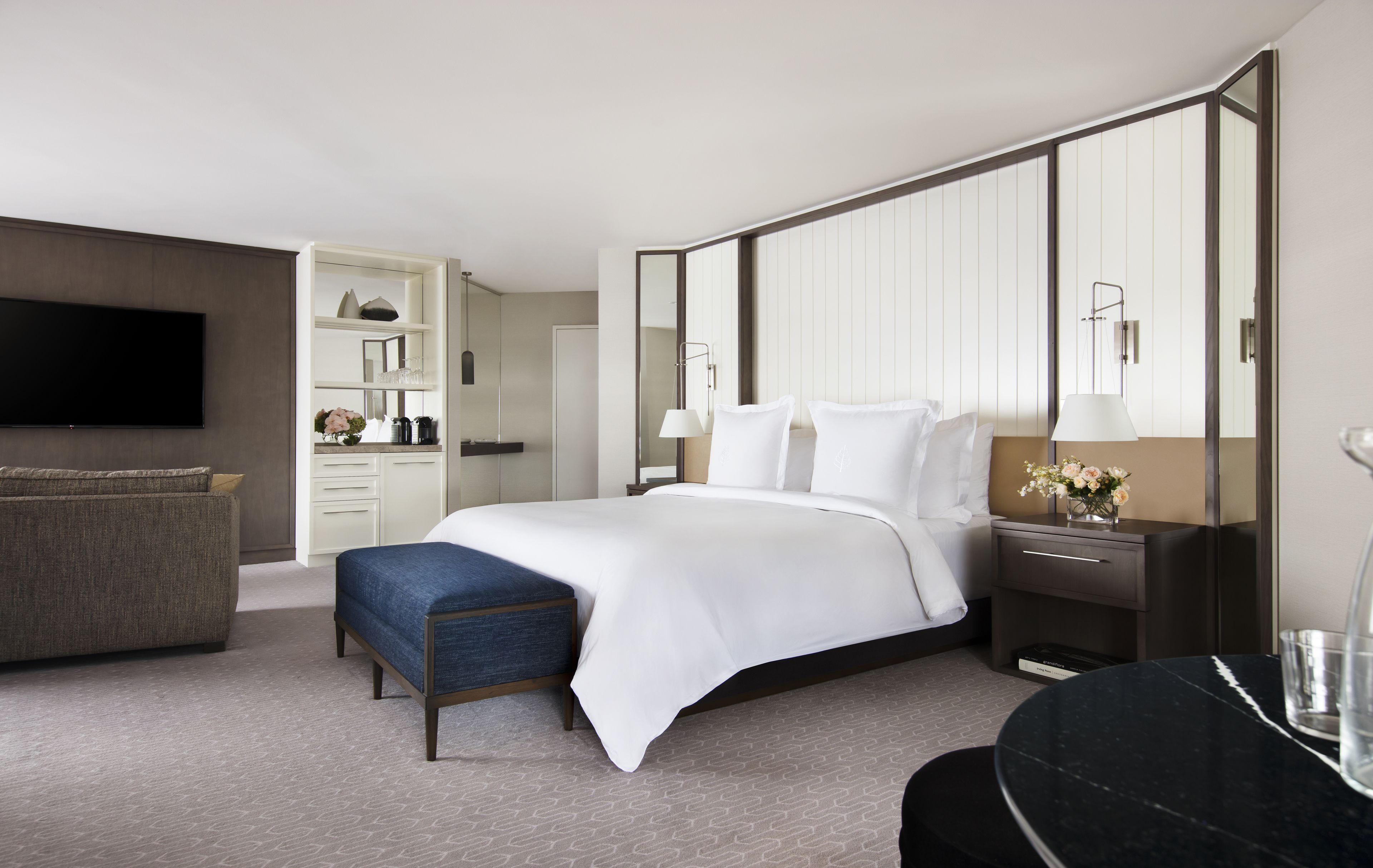 A modern hotel room with a large bed, a seating area with a sofa and ottoman, a flat-screen TV, a vanity with a mirror, and a built-in wardrobe.