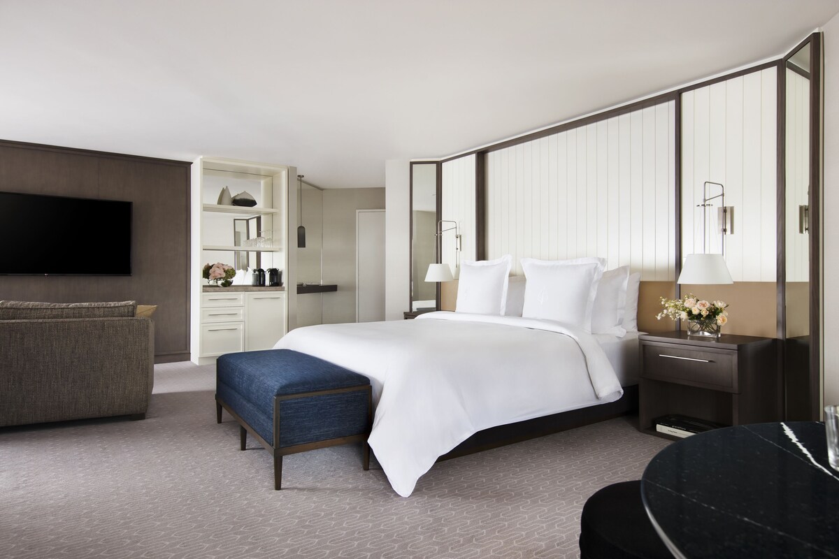 A modern hotel room with a large bed, a seating area with a sofa and ottoman, a flat-screen TV, a vanity with a mirror, and a built-in wardrobe.