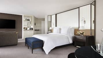 A modern hotel room with a large bed, a seating area with a sofa and ottoman, a flat-screen TV, a vanity with a mirror, and a built-in wardrobe.