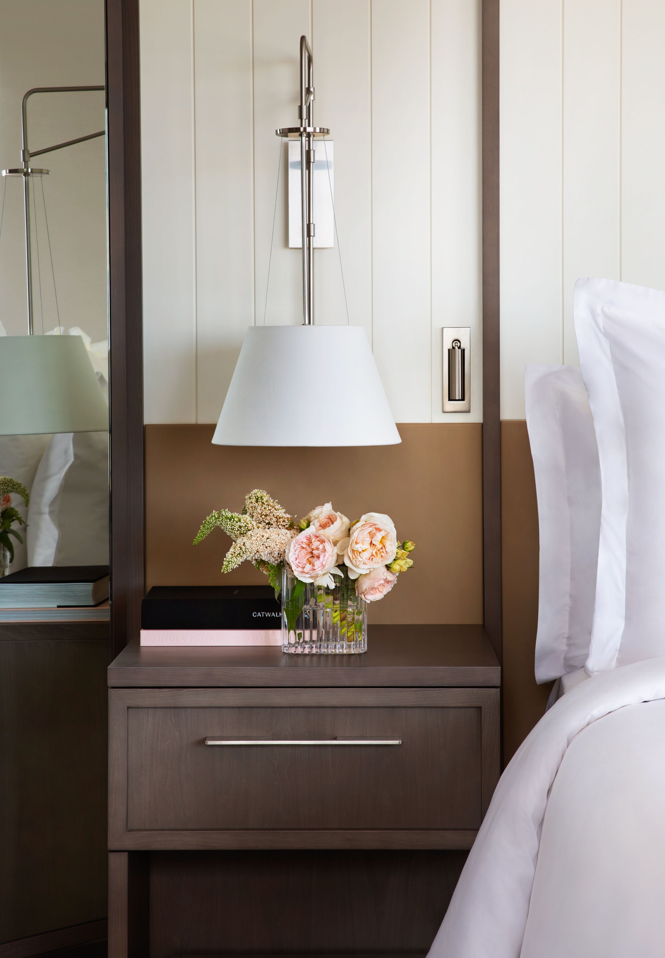 Una habitación de hotel con una mesita de noche, una lámpara, un jarrón con flores y una cama con ropa blanca.