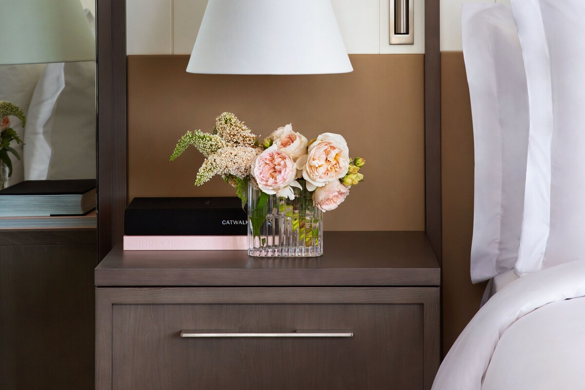 A hotel room with a bedside table, a lamp, a vase with flowers, and a bed with white linens.