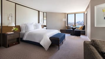 A modern hotel room with a large bed, a seating area with a view, and a cityscape visible through the window.