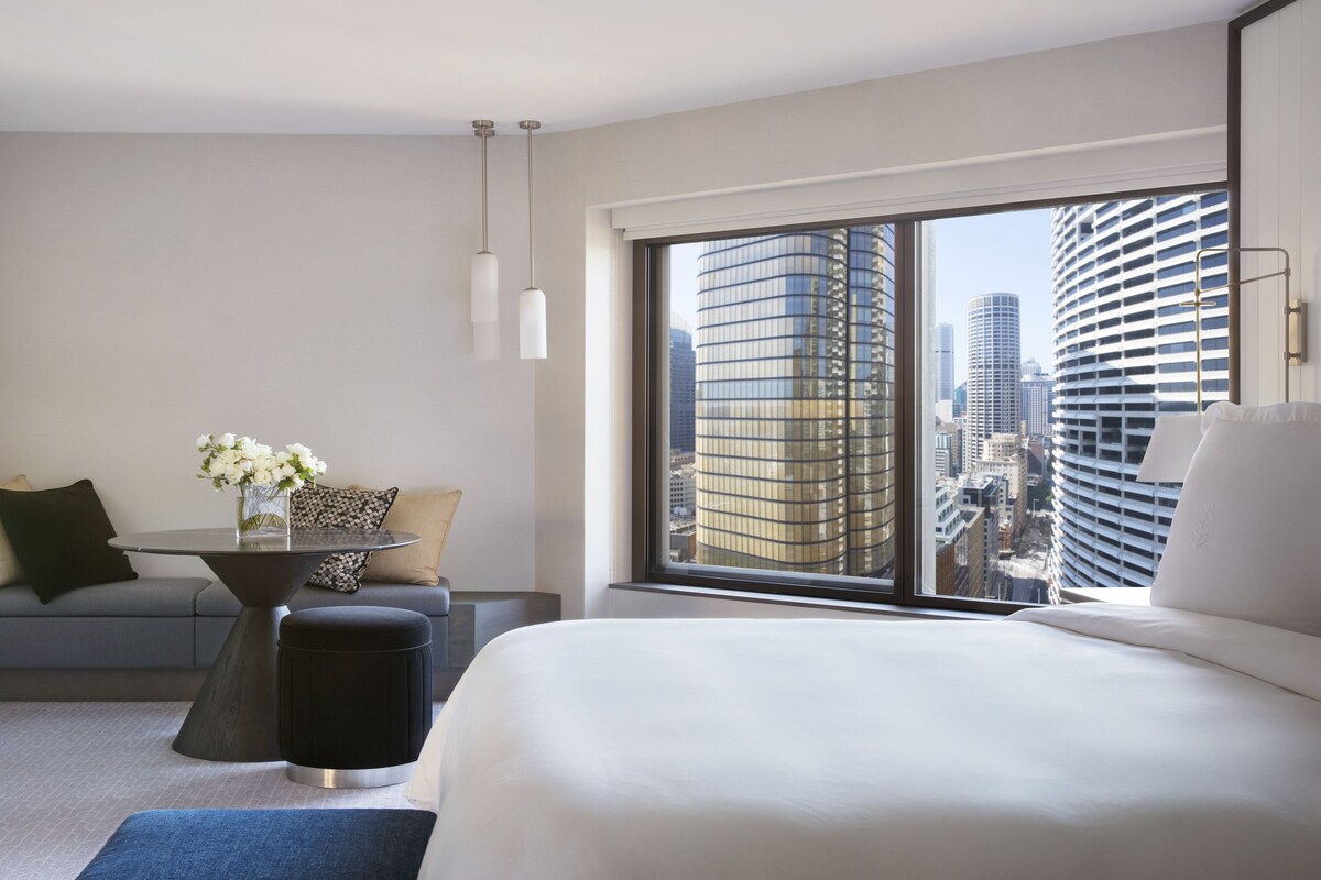 A modern hotel room with a large bed, a sofa, a small table with flowers, and a view of city buildings.