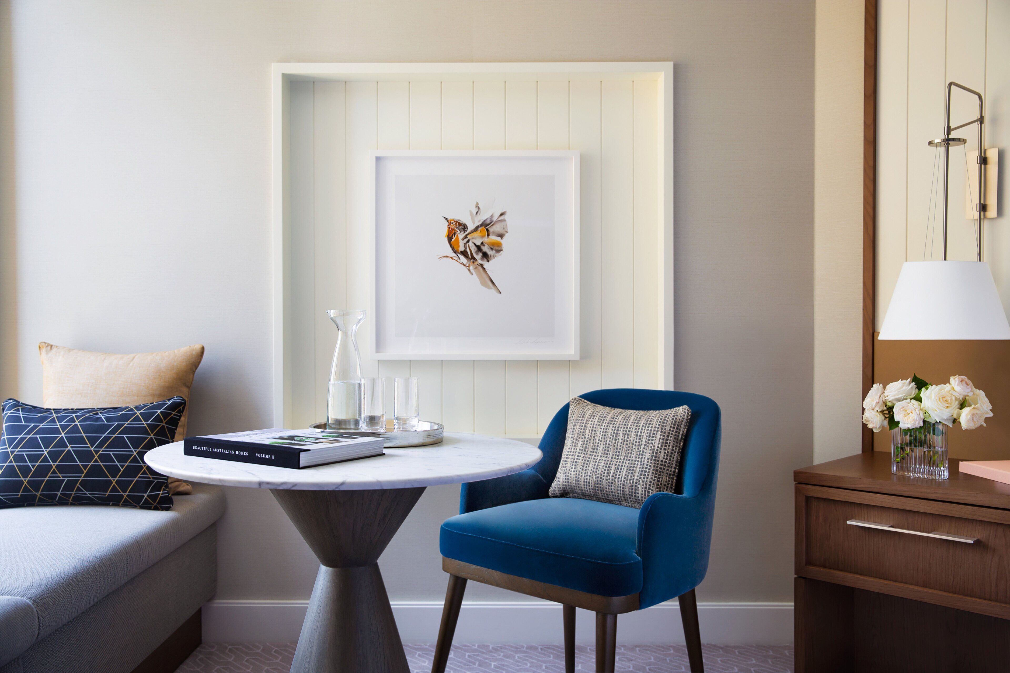 A modern room with a blue armchair, a small round table with a book and glasses, a framed artwork on the wall, and a nightstand with a lamp and vase of flowers.