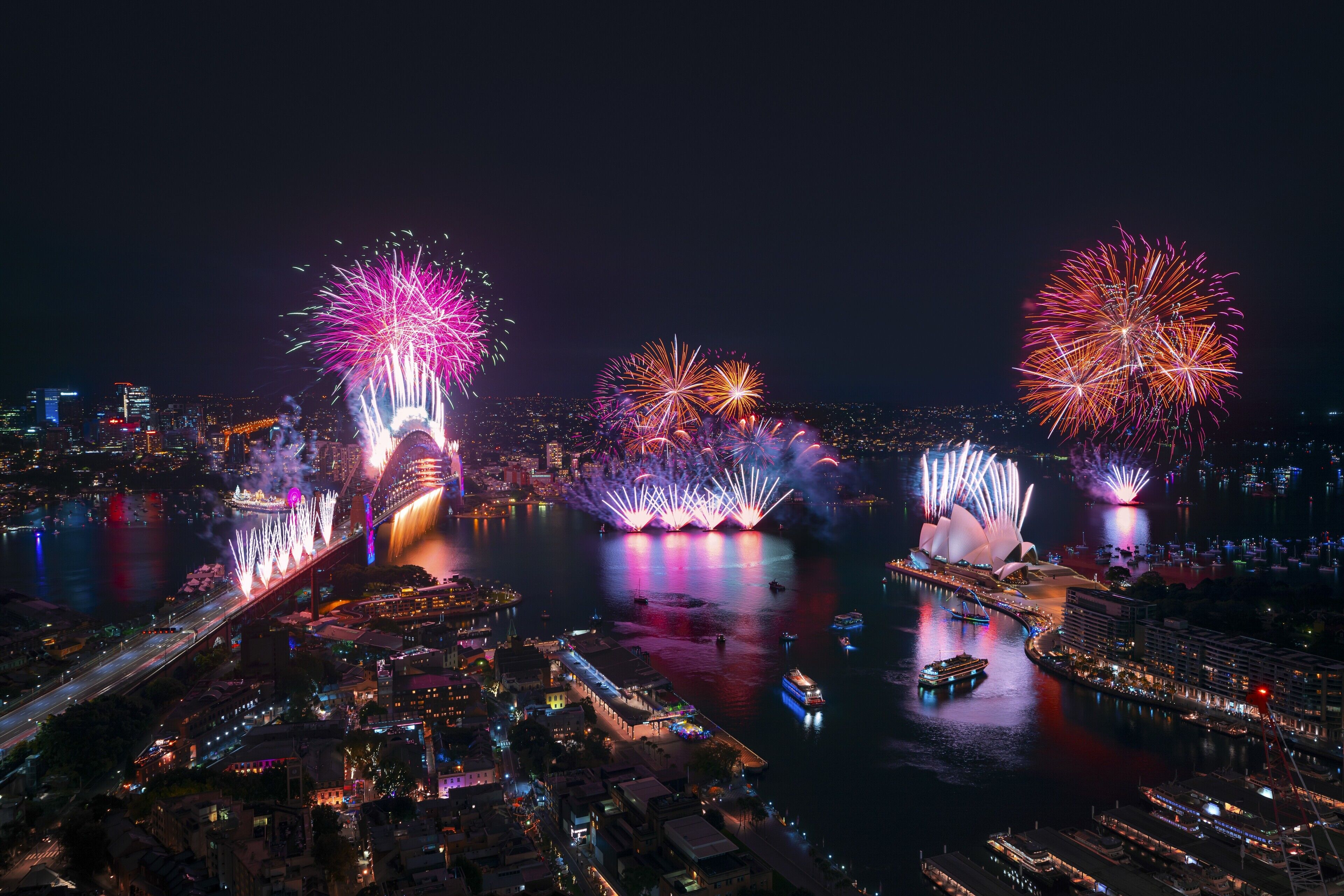 A nighttime cityscape with fireworks over a river, featuring illuminated buildings and a bridge.