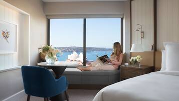 A hotel room with a large window, a bed, a bedside table with a lamp, a chair, and a small table with a vase of flowers.