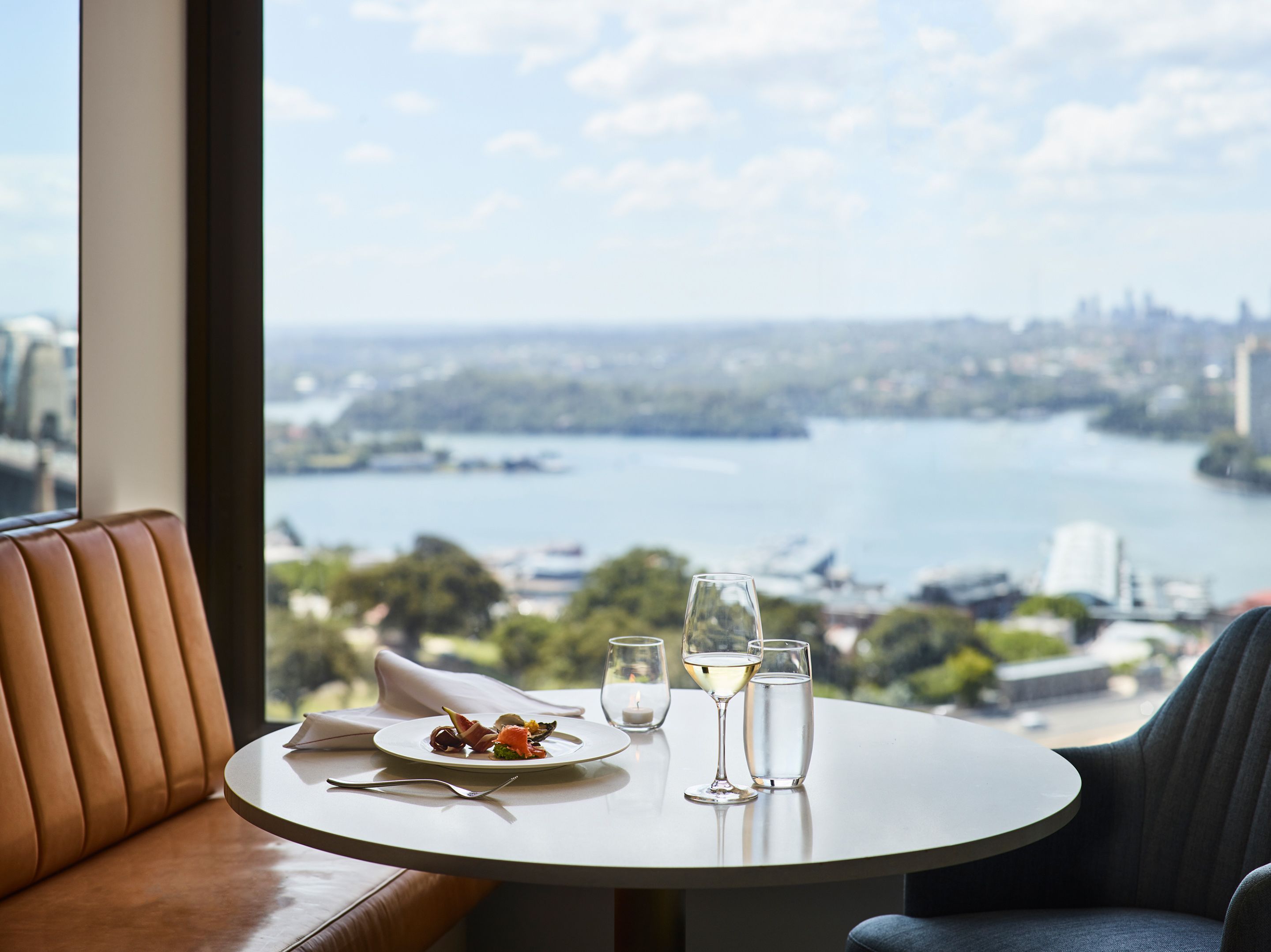 A dining table with a plate of food and two glasses of white wine, set against a scenic view of a river and cityscape.