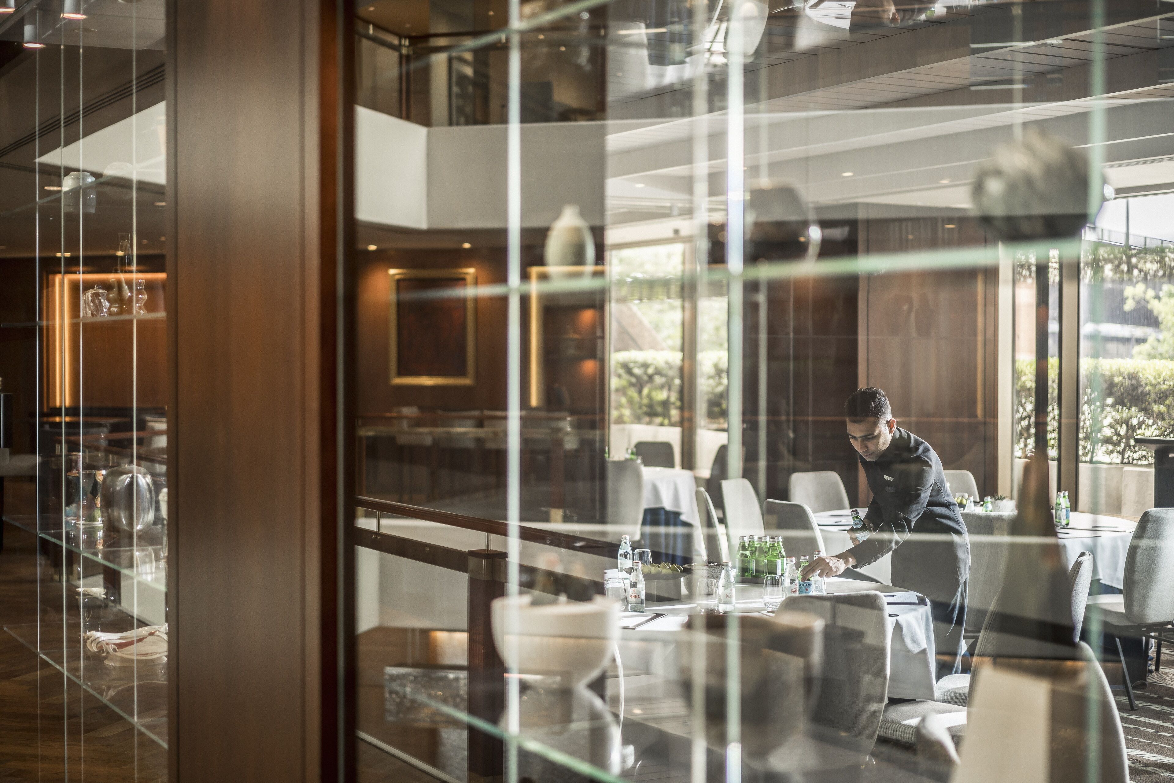A modern dining area with glass partitions, a waiter attending to guests, and a view of the outdoors.