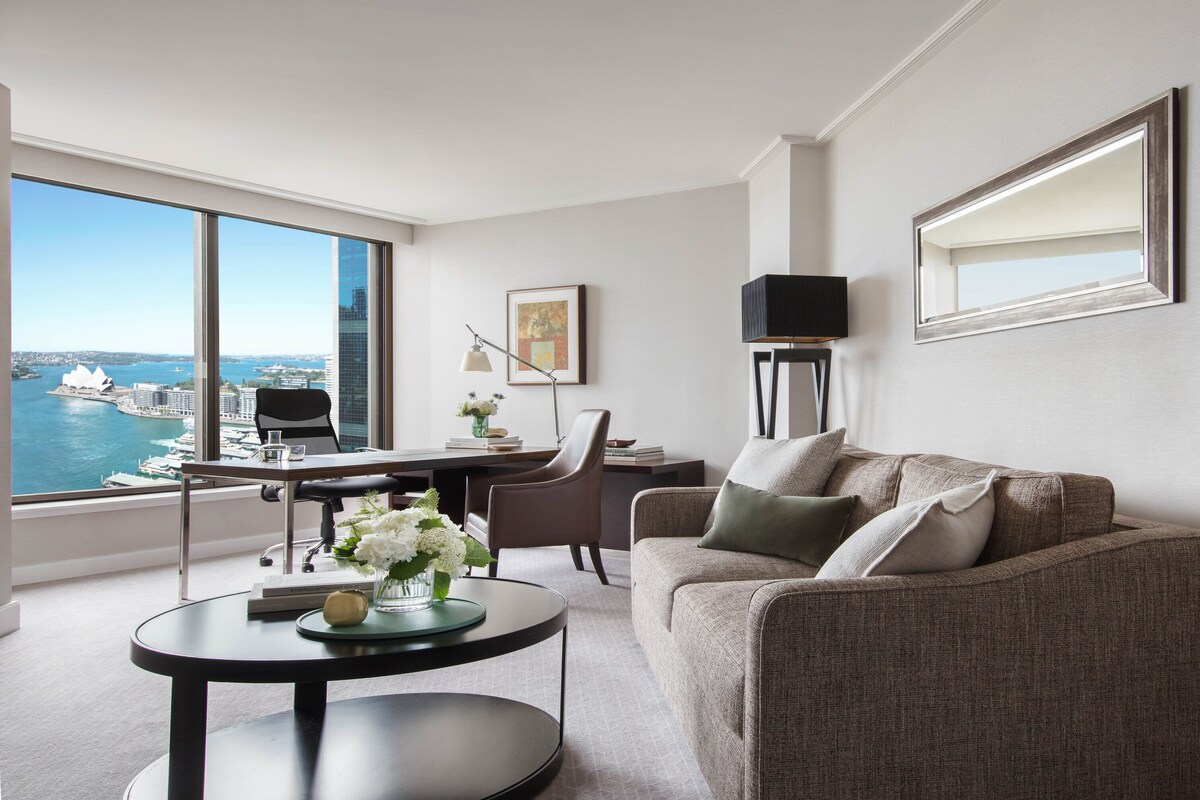 A modern living room with a view of a cityscape and water. It includes a sofa, a coffee table, a desk, and a chair.