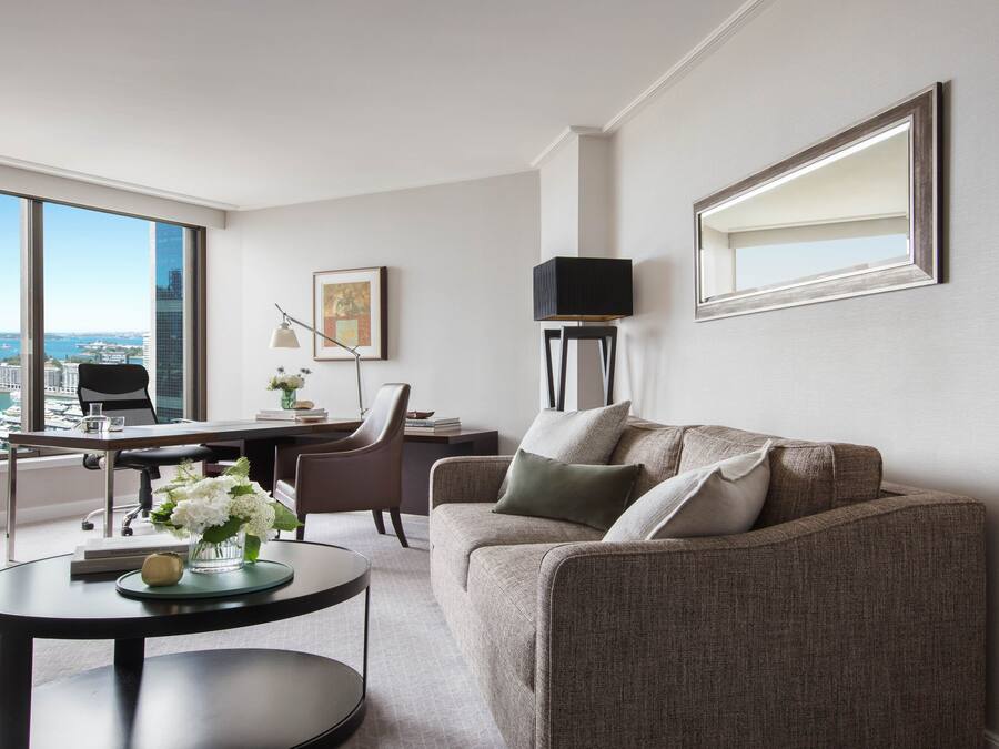 A modern living room with a view of a cityscape and water. It includes a sofa, a coffee table, a desk, and a chair.