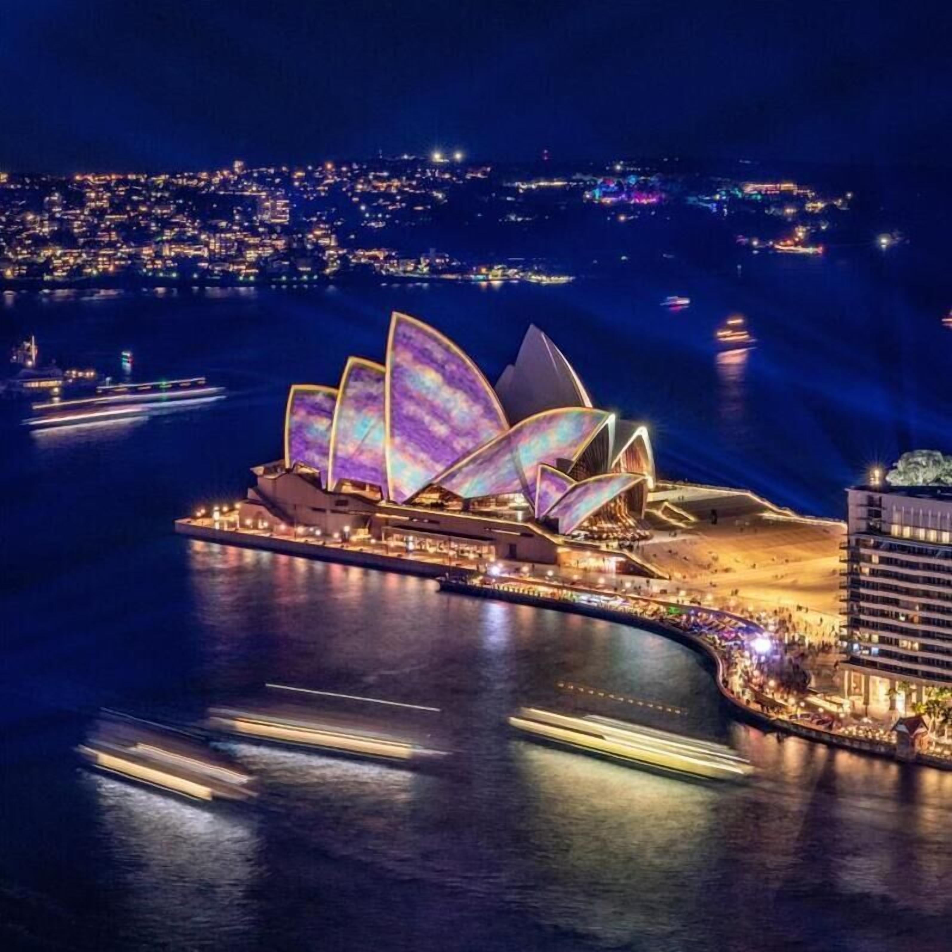 Une vue nocturne de l'Opéra de Sydney, reconnaissable à son architecture originale évoquant des voiles, située au bord de l'eau, avec la silhouette de la ville en arrière-plan.