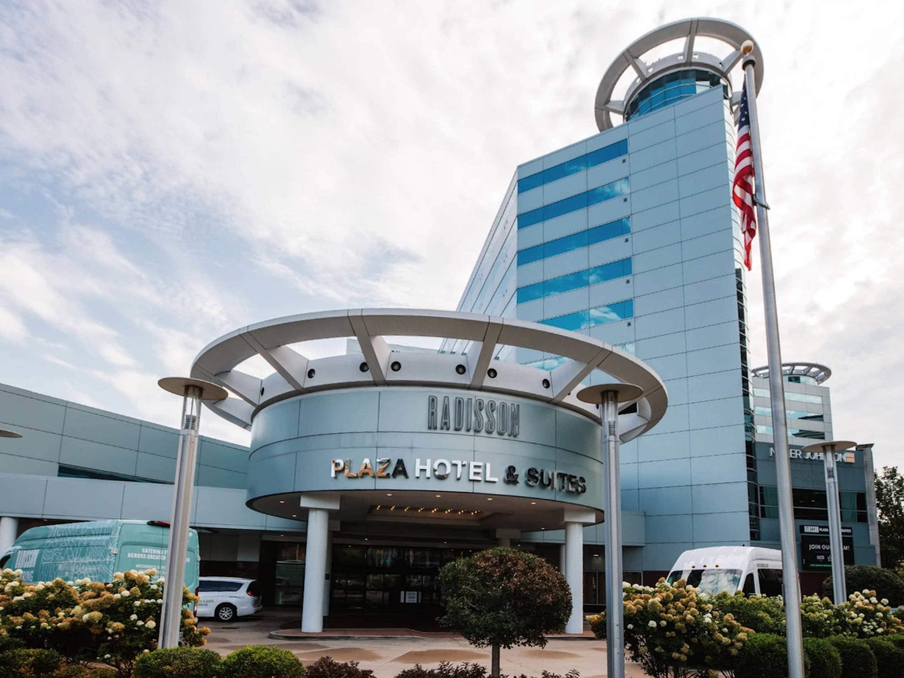 A modern hotel with a curved entrance, a prominent American flag, and a sign reading 'RADISSON PLAZA HOTEL & SUITES'.