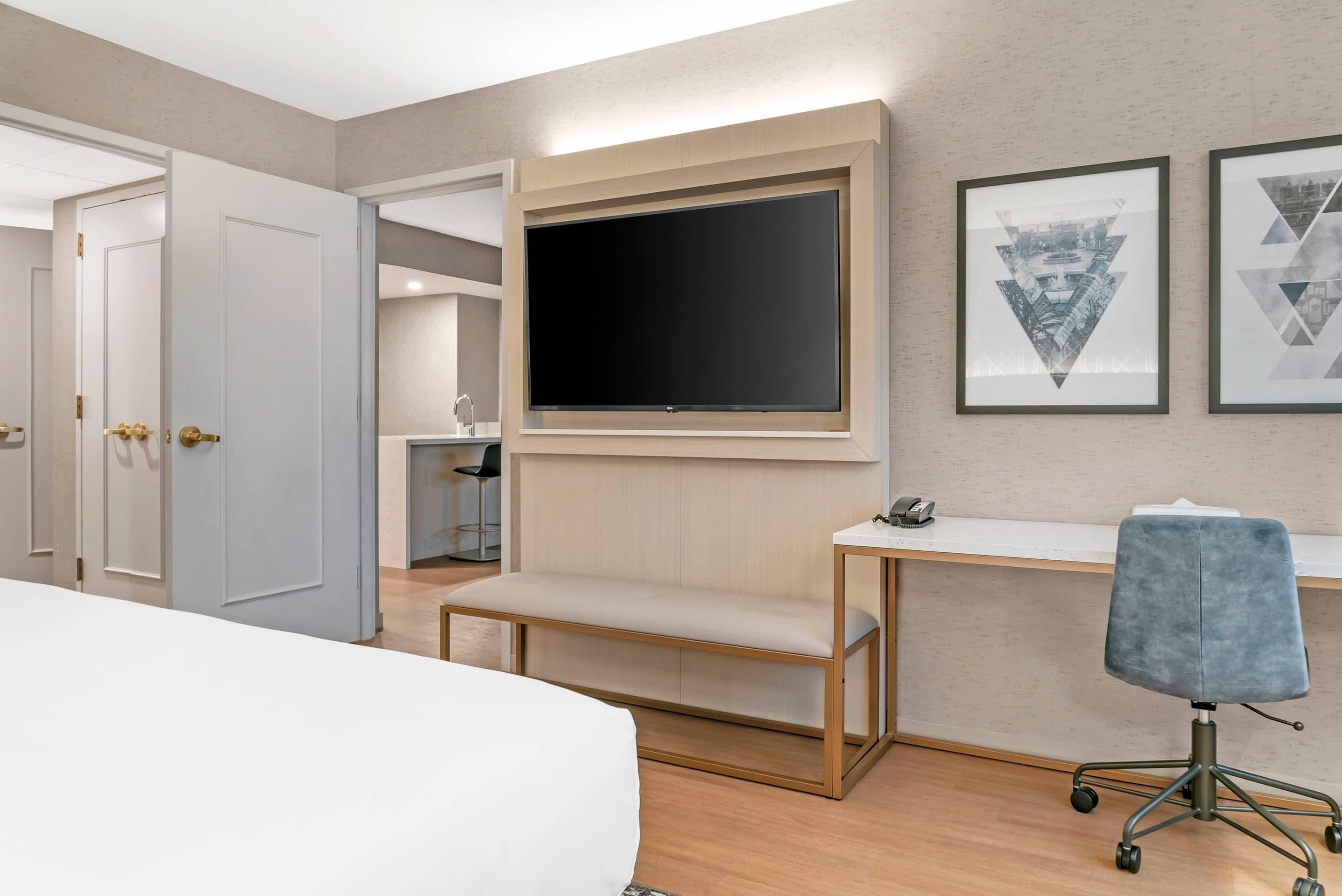 A hotel room with a bed, a desk with a chair, a mounted TV, and artwork on the wall.