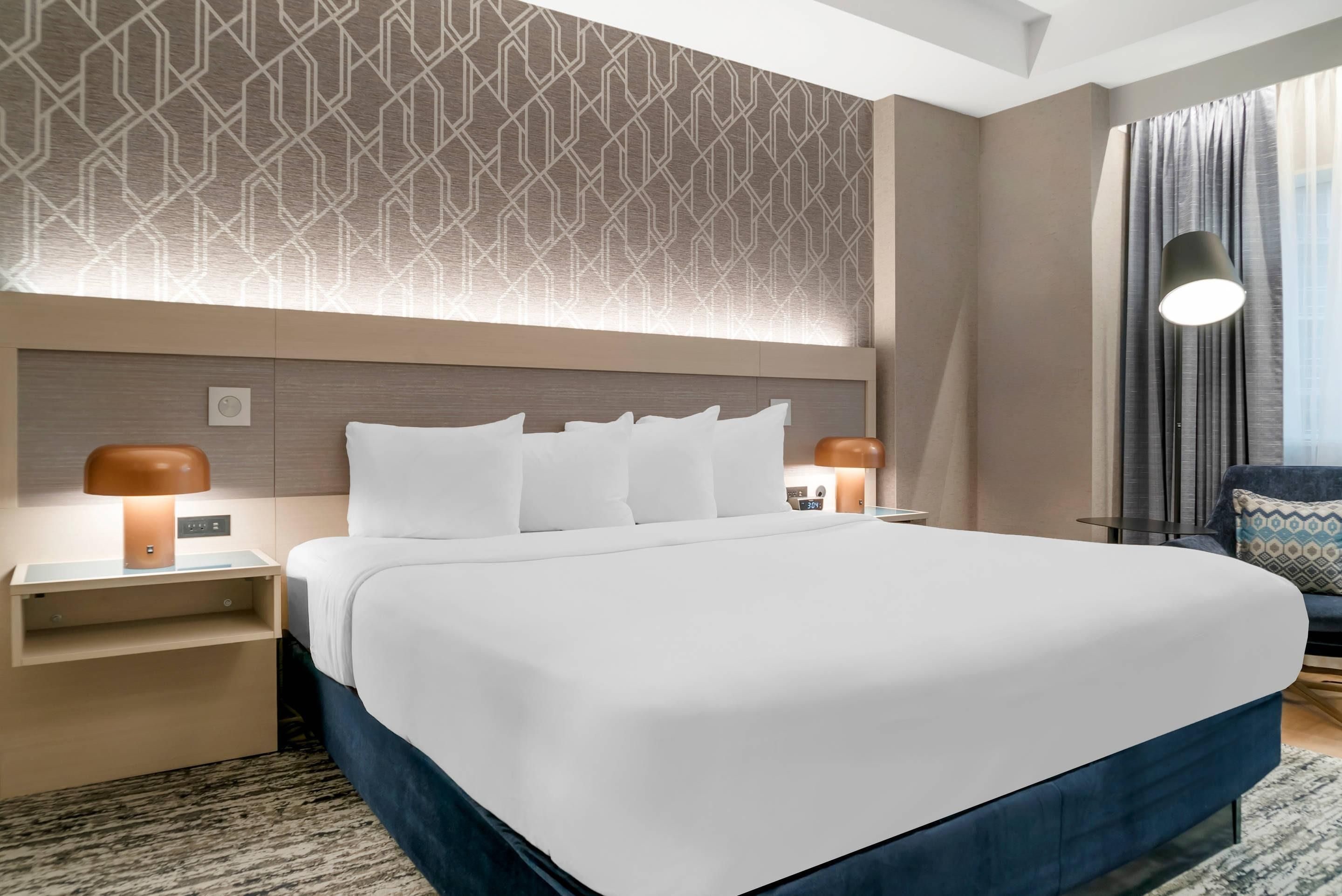 Un moderno cuarto de hotel con una cama grande, dos mesitas de noche con lámparas, un cabecero con diseño geométrico y vistas a la ciudad desde la ventana.
