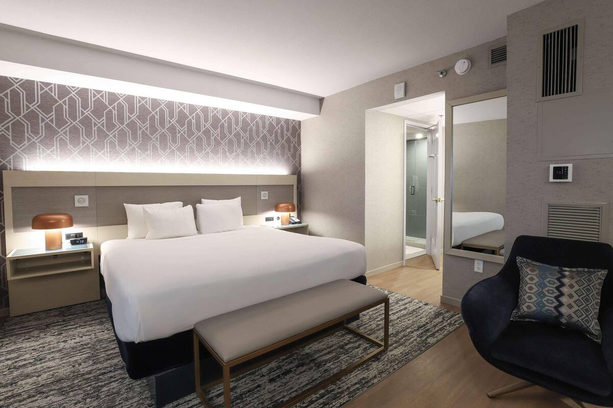 Una habitación de hotel moderna con una cama grande, mesitas de noche, un banco, una silla y un papel tapiz estampado.