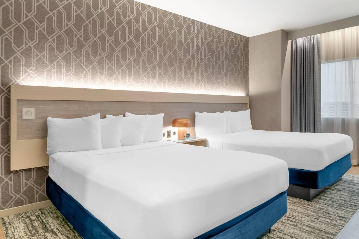 Habitación de hotel con dos camas, cabecero con iluminación integrada, mesita de noche con lámpara y un ventanal con cortinas.