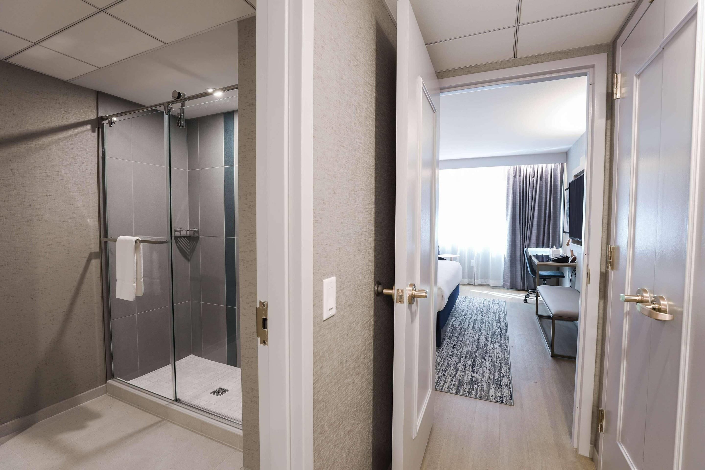 A hotel room with a glass-enclosed shower, a bed with a view, a desk, and a television.