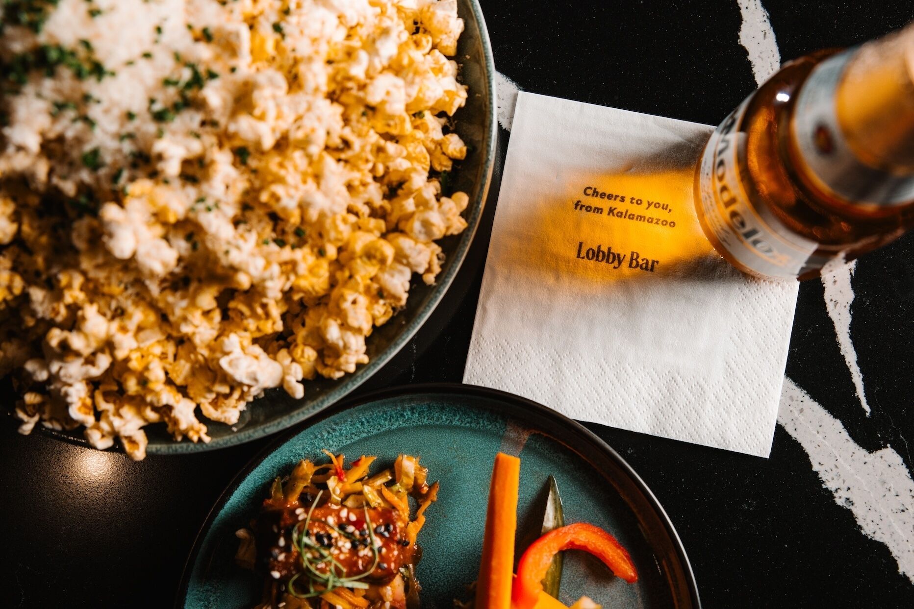 A bowl of popcorn, a bottle of beer, and a plate with vegetables on a table.
