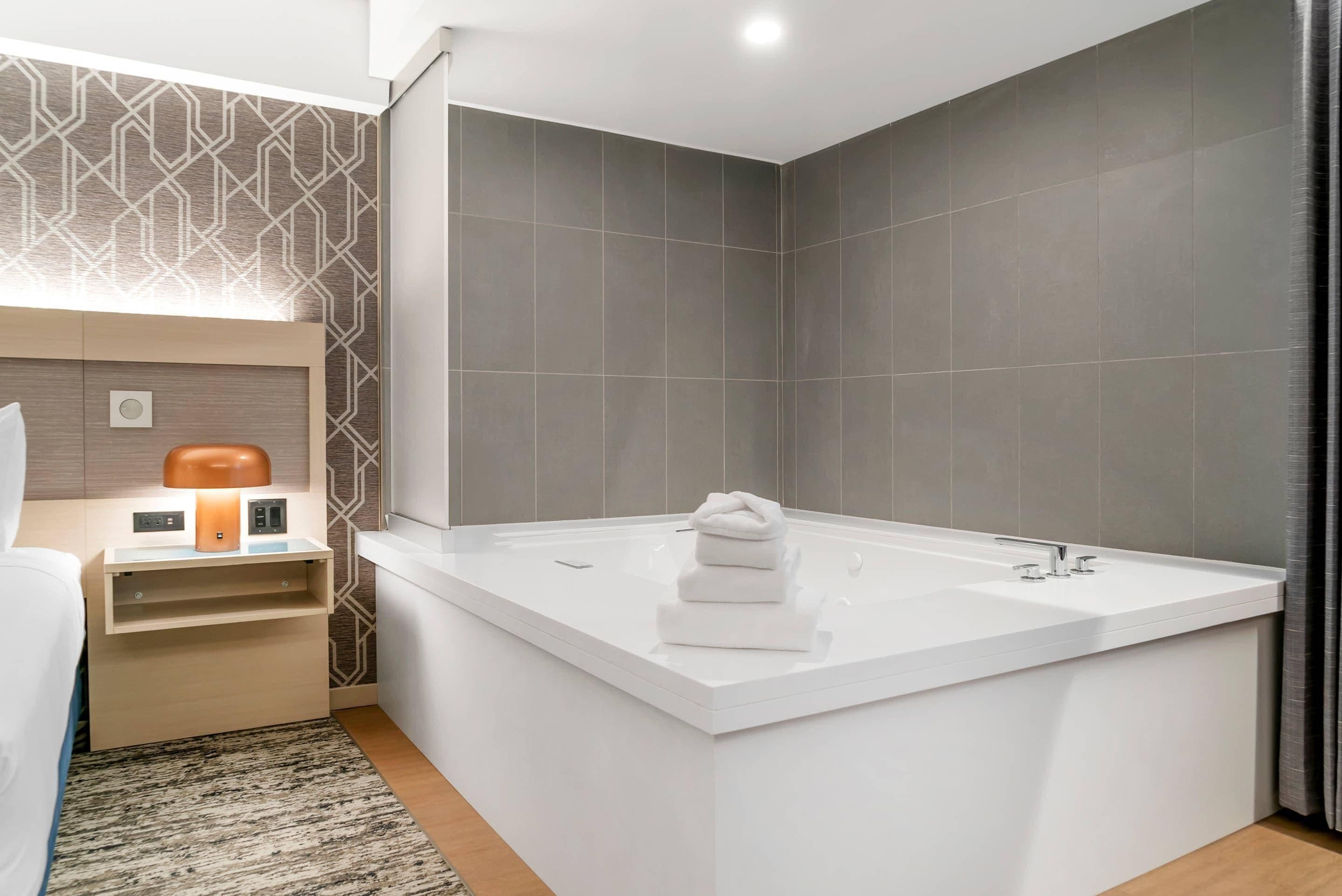 A modern bathroom with a large white bathtub, a wooden nightstand with a lamp, and a patterned wall.