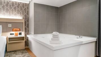A modern bathroom with a large white bathtub, a wooden nightstand with a lamp, and a patterned wall.