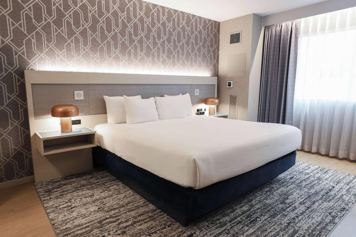 Una habitación de hotel moderna con una cama grande, una mesita de noche con lámpara y un papel tapiz estampado.