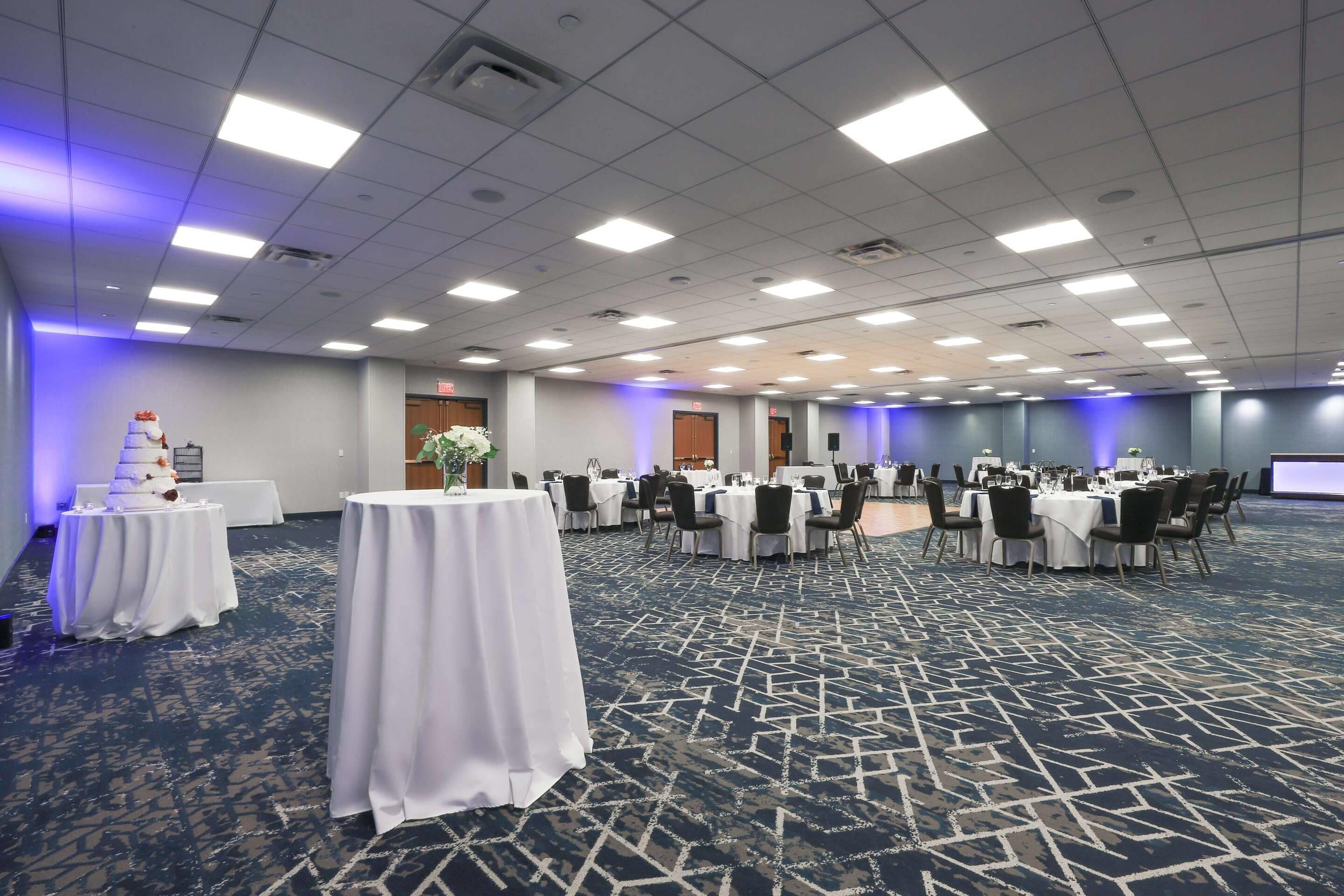 Un salón de conferencias con mesas dispuestas para una reunión, un pastel sobre una de ellas y una pantalla de proyector al fondo.