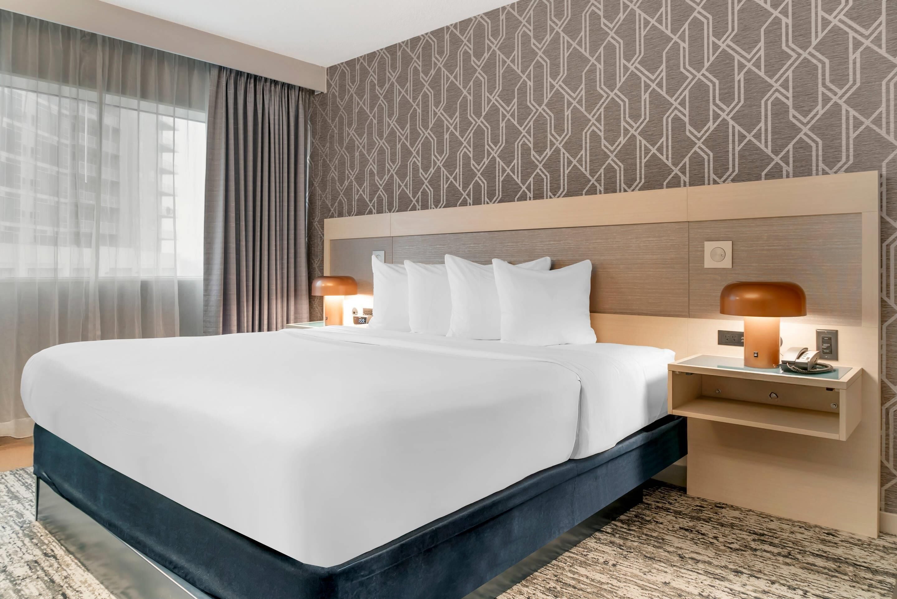 Habitación de hotel con una cama grande, dos mesitas de noche con lámparas, un cabecero y una pared con estampado.