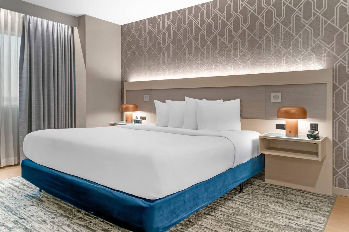 Una habitación de hotel moderna con una cama grande, dos mesitas de noche con lámparas, un papel tapiz decorativo y un amplio ventanal con cortinas.