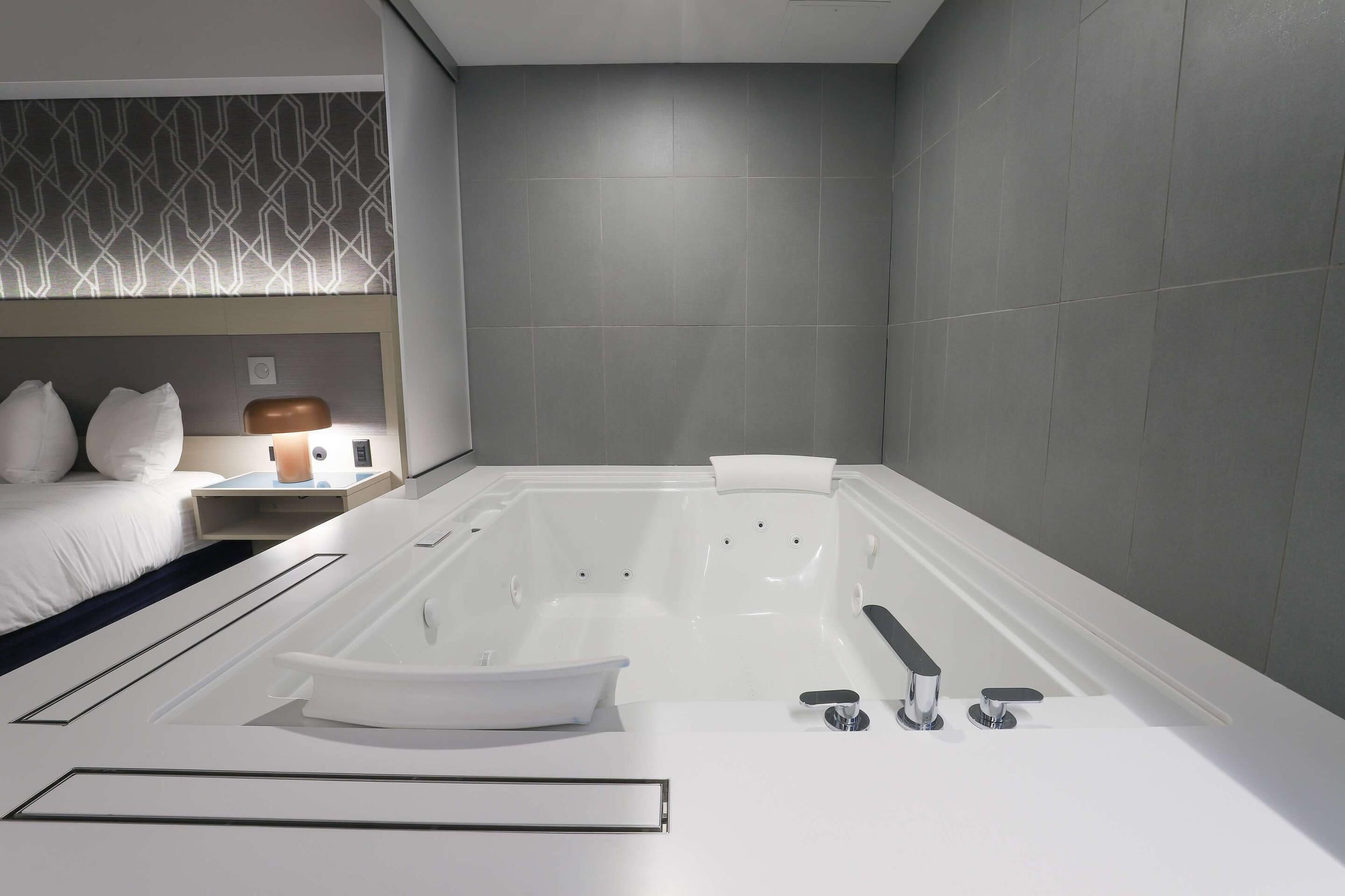 A modern bathroom with a large bathtub, a white sink, and a bedside table with a lamp.