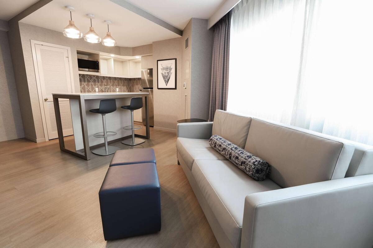 Una sala de estar moderna con sofá y reposapiés, un comedor con taburetes de bar, y una cocina con microondas y refrigerador.