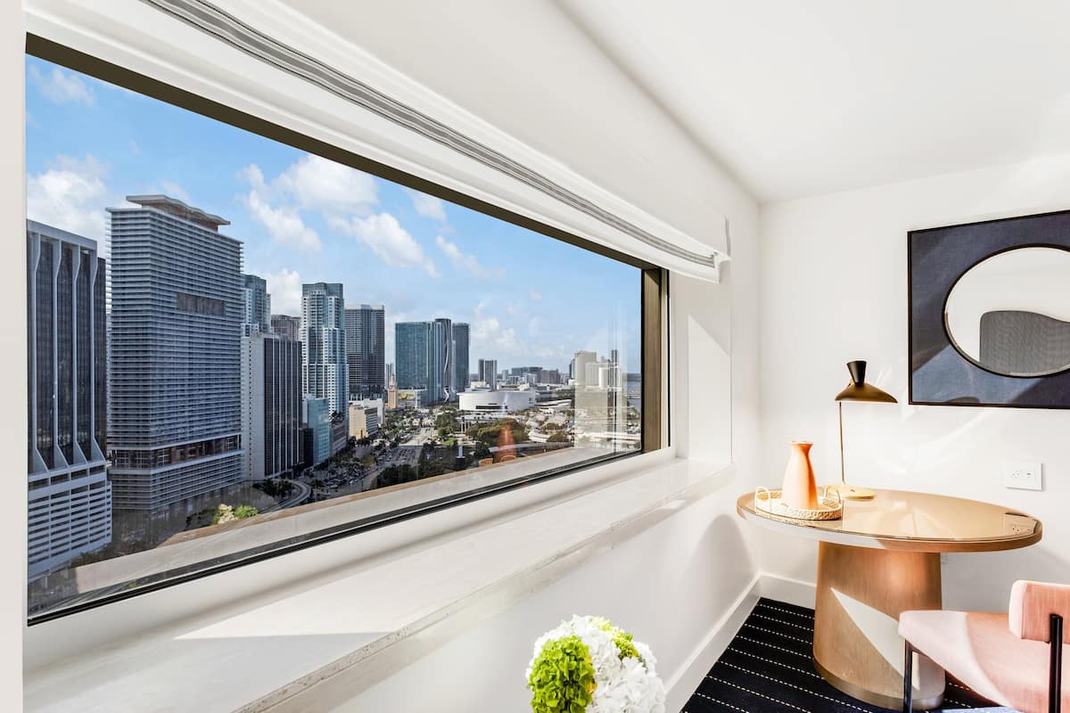 A modern hotel room with a large window offering a city view, a round table with a lamp, and a chair.