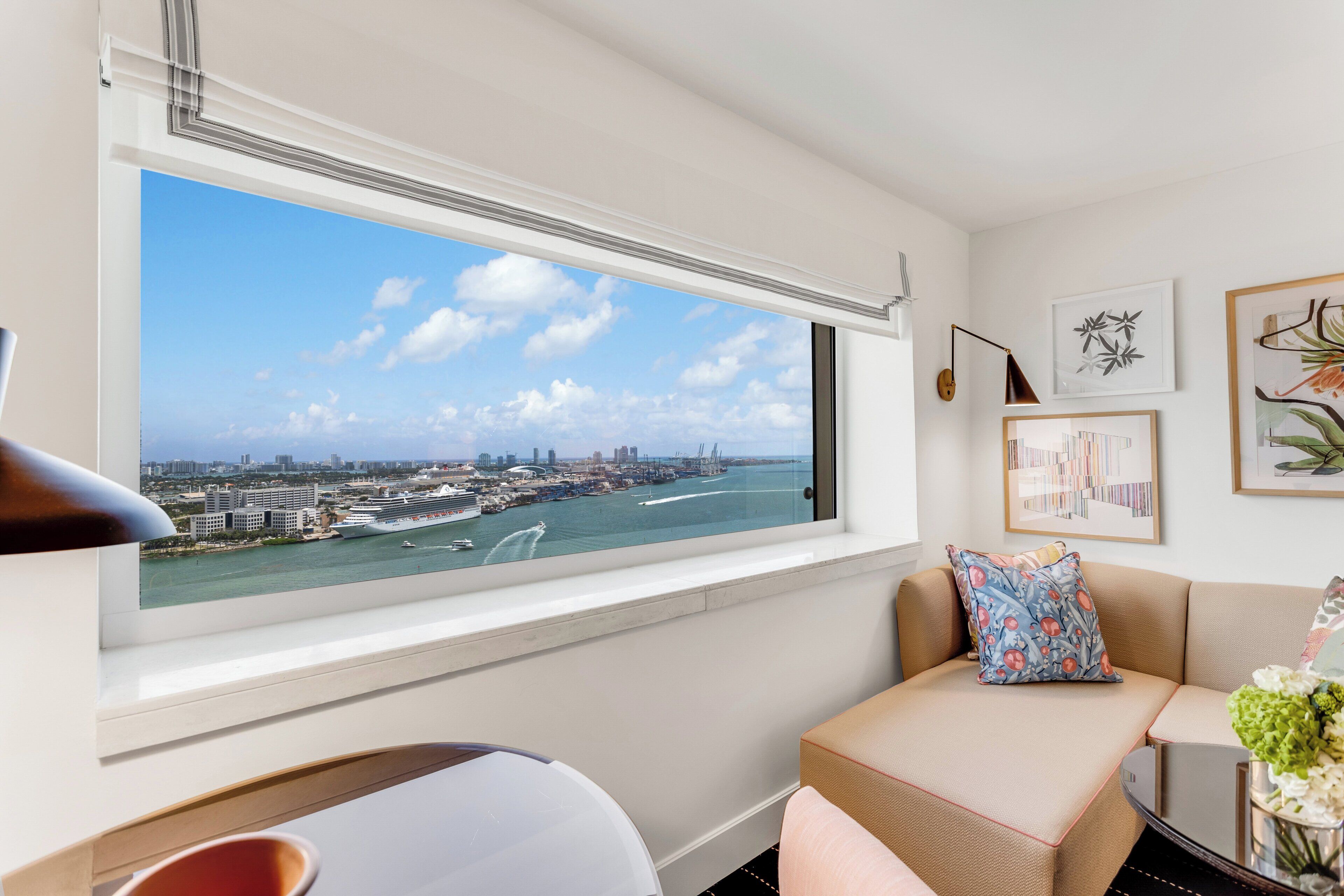 A room with a large window overlooking a marina and city skyline, a sofa with patterned cushions, and a wall-mounted lamp.