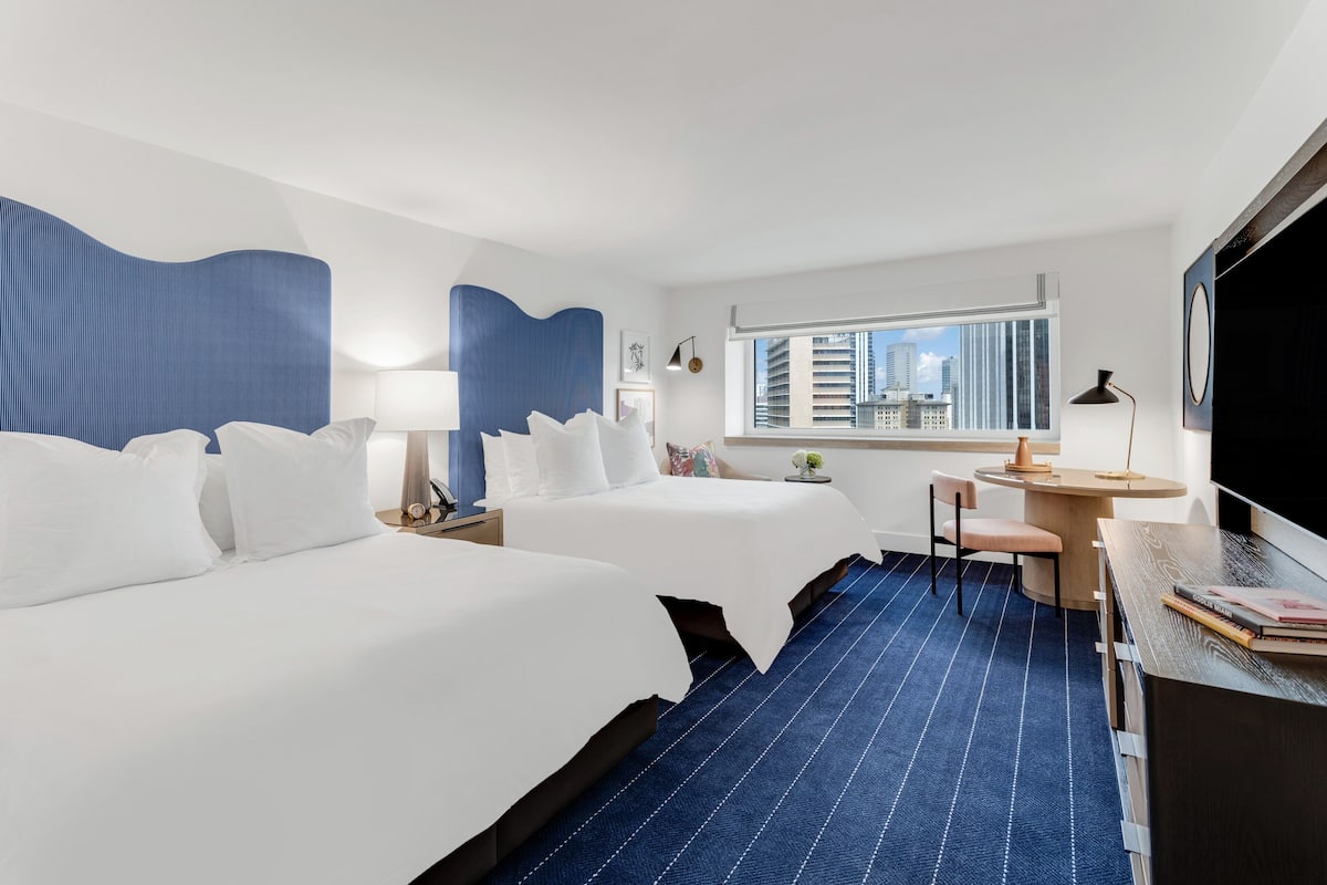 A hotel room with two beds, a desk, a chair, a window with a city view, and a large TV.
